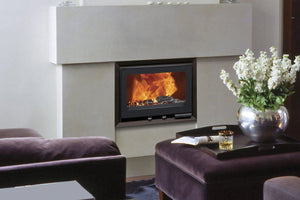 Fondis - V80L - 11.5kW Inset Wood Stove with flames in a lounging area with an armchair and an ottoman